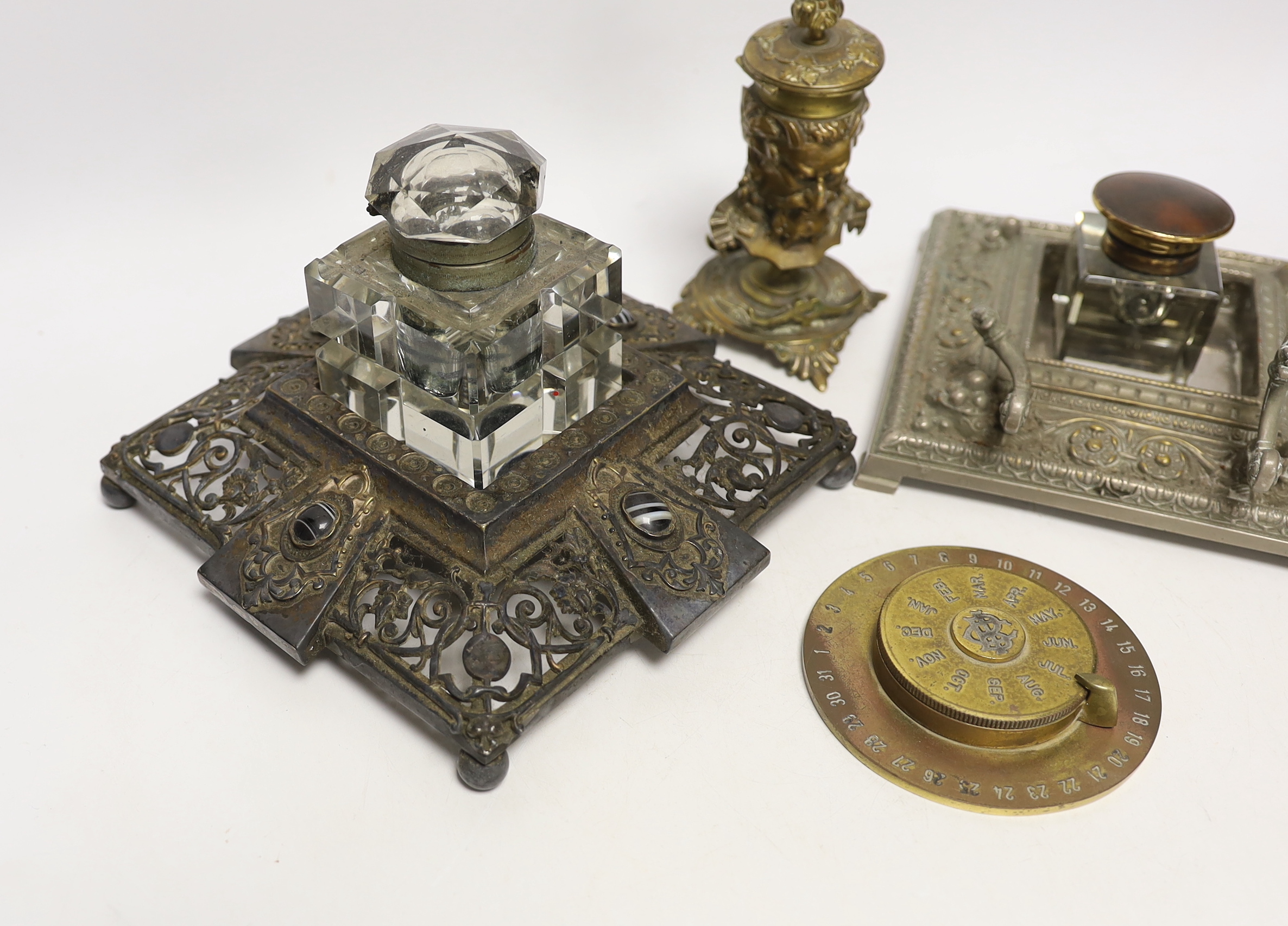 Three cast metal and cut glass ink stands, including a bronze inkwell in the form of the head of Bacus, and a brass calendar display from the Banca Commerciale Italiana, London Branch, tallest 13cm